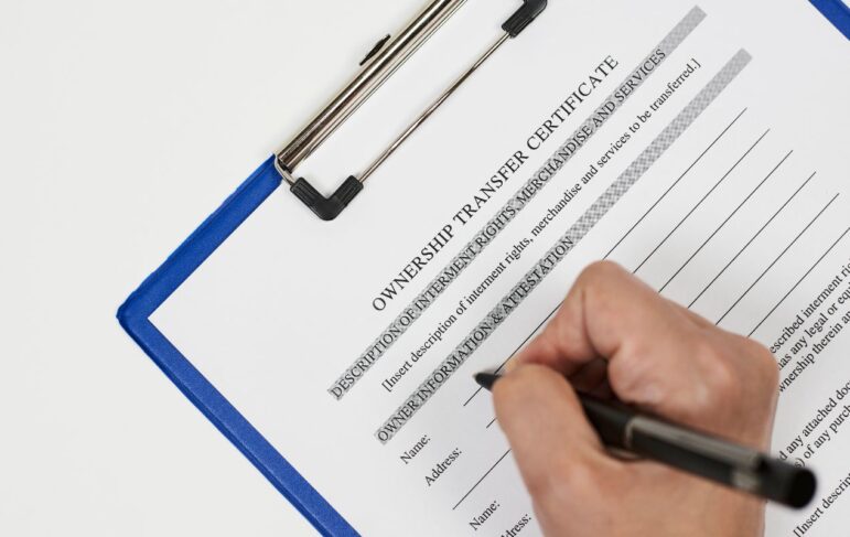 person's hand holding a black pen while filling out a paper that reads "ownership transfer certificate" on a blue clipboard